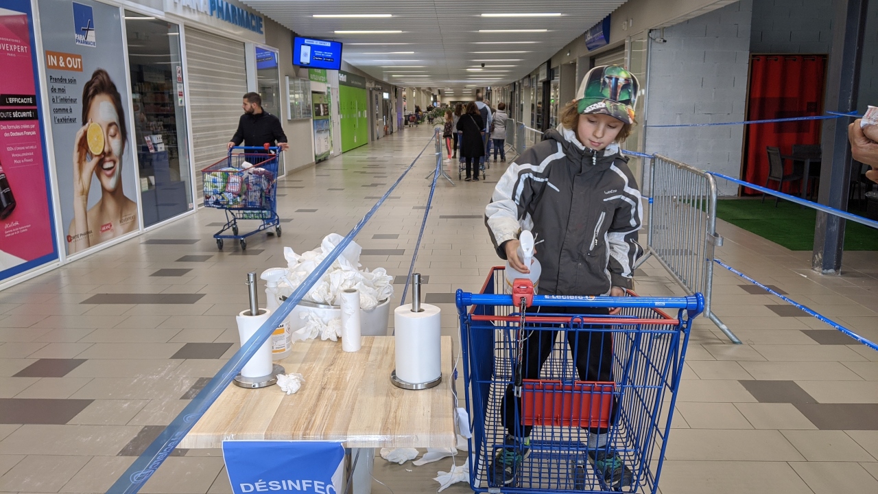 Ein Junge desinfiziert mit einem Reinigungsspray einen Einkaufswagen in einem Supermarkt. Dazu hat er sich in den Einkaufswagen begeben. Im Hintergrund ist eine Warteschlange mit Menschen, die den Supermarkt betreten möchten. 