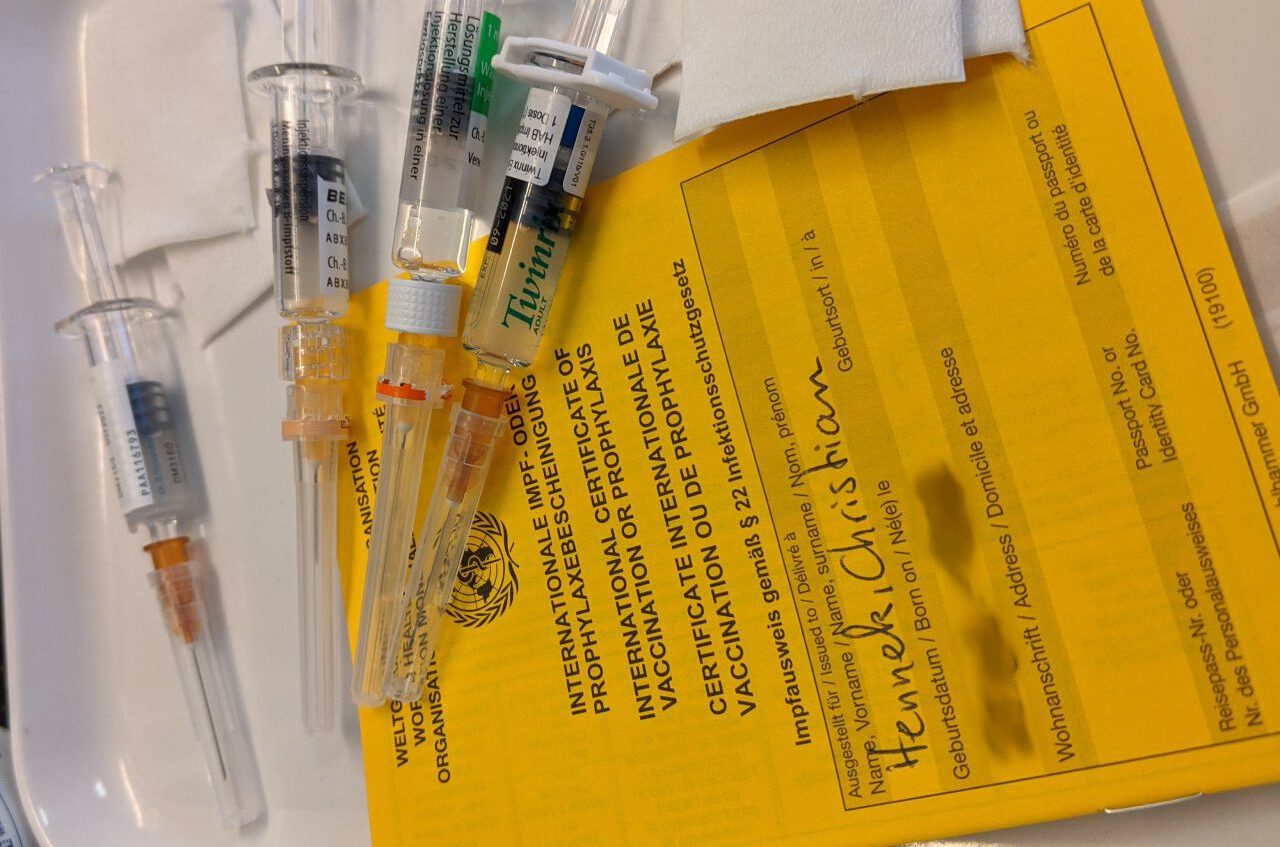 vier vorbereitete spritzen mit unterschiedlichen Impffstoffen liegen auf einem Tablet bereit, ausserdem ist ein oranger Impfpass abgebildet