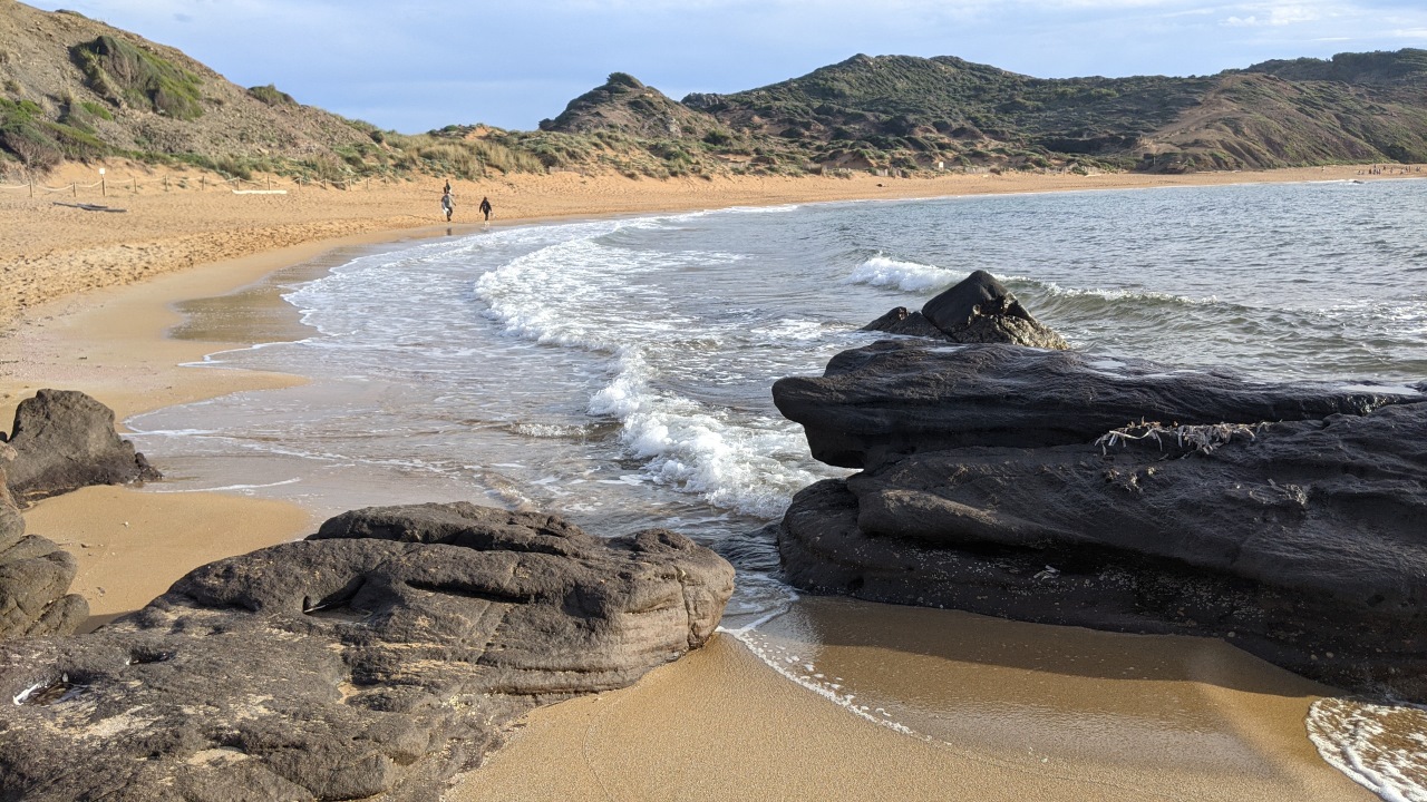 Das Bild zeigt den Strand Platja de Cavalleria im Norden Menorcas mit Felsen im Vordergrund und ein paar Spaziergängern