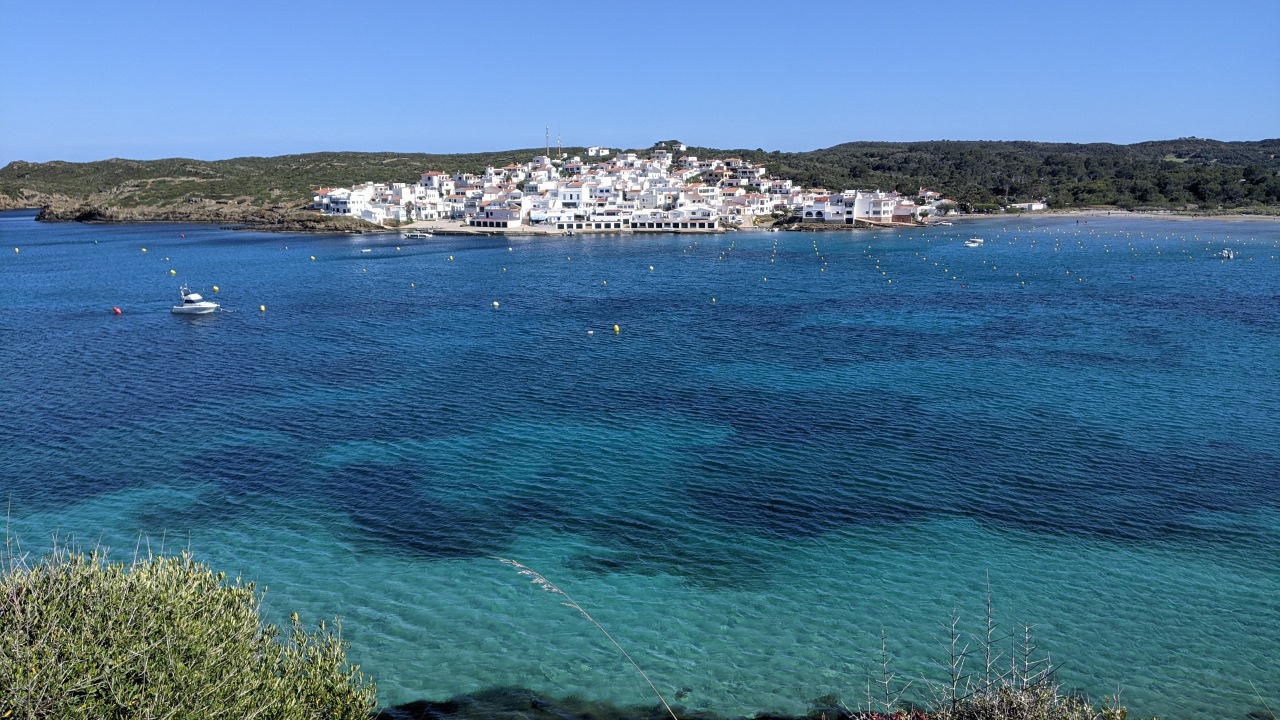 Der Ort Es Grau auf Menorca, im Vordergrund ist türkis blaues Wasser 