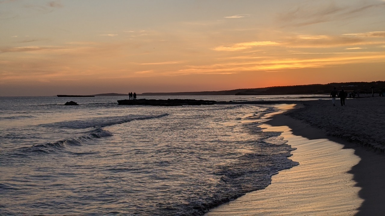 Sonnenuntergang auf Menorca, Menschen blicken von einem Felsvorsprung aufs Meer während die Wellen an den Strand laufen