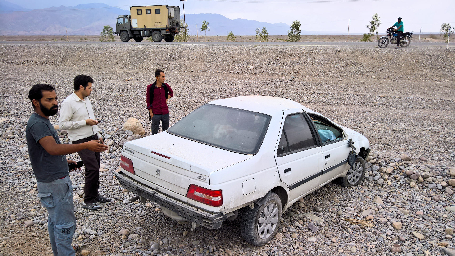 Ein Peugeot 405 steht nach einem Unfall zerbeult am Rand einer Strasse. Drei Männer betrachten das beschädigte Auto. 