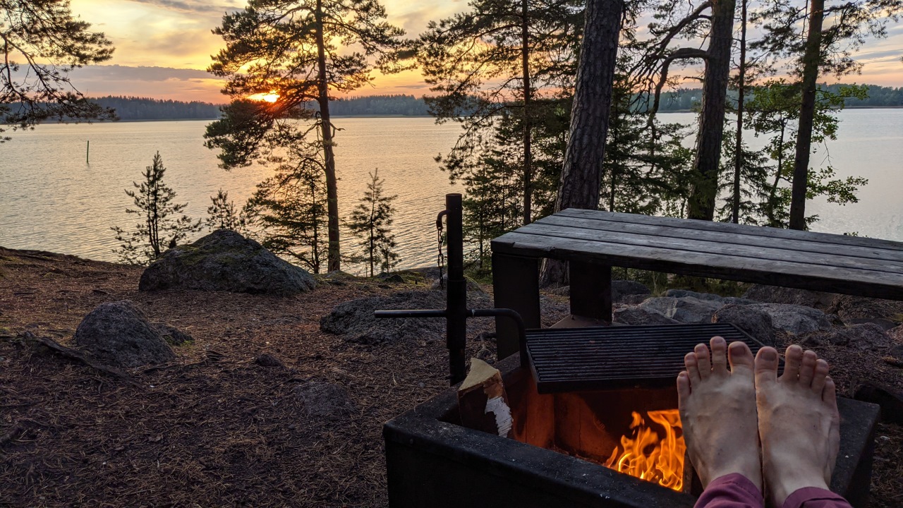 Ein finnischer Rastplatz mit Feuerstelle und Blick auf einen See. Ich wärme mit am Feuer die nackten Füsse. 