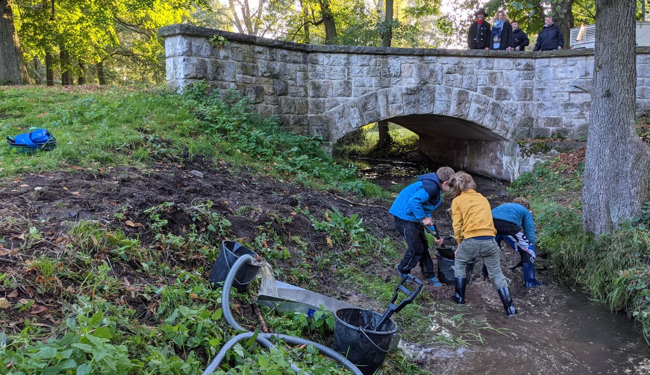 Drei Kinder graben Sand aus einem kleinen Bach, um damit eine Goldwaschrinne zu füllen. Im Hintergrund sind Zuschauer auf einer Stein Brücke. 