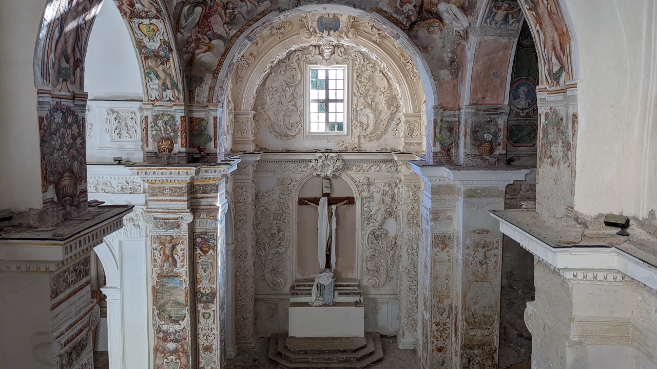 Innenansicht der Kirche Santa Maria delle grazie. Der Innenraum ist reicht mit Stuck Arbeiten und farbigen Malereien verziert. Jesus Christus ist mit einem Laken verhüllt. Es sind Wasserschäden erkennbar, Fensterscheiben fehlen und es liegt viel Staub. 