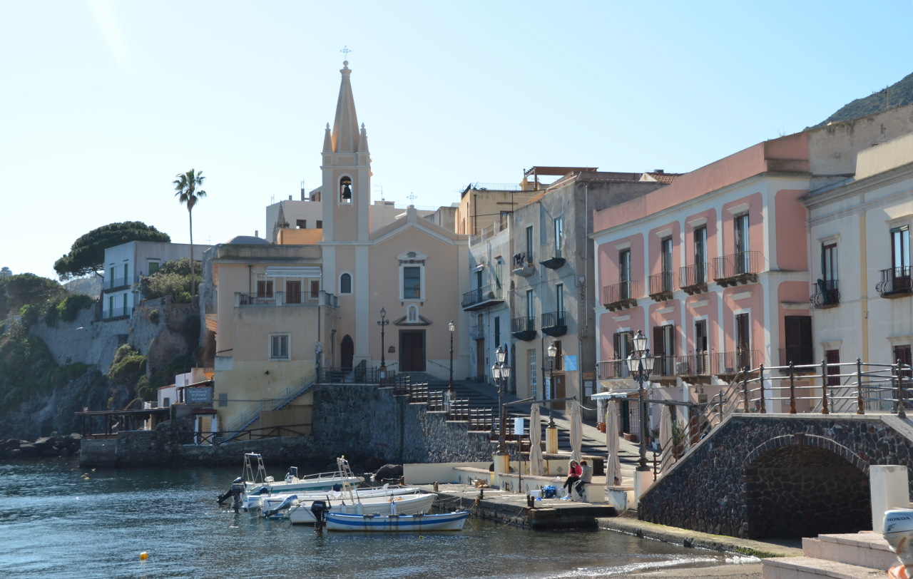 Fischerhafen von Lipari. Auf dem Wasser schaukeln ein paar kleine Boote, im Hintergrund ist eine Kirche zu der ein Fußweg ansteigt. 
