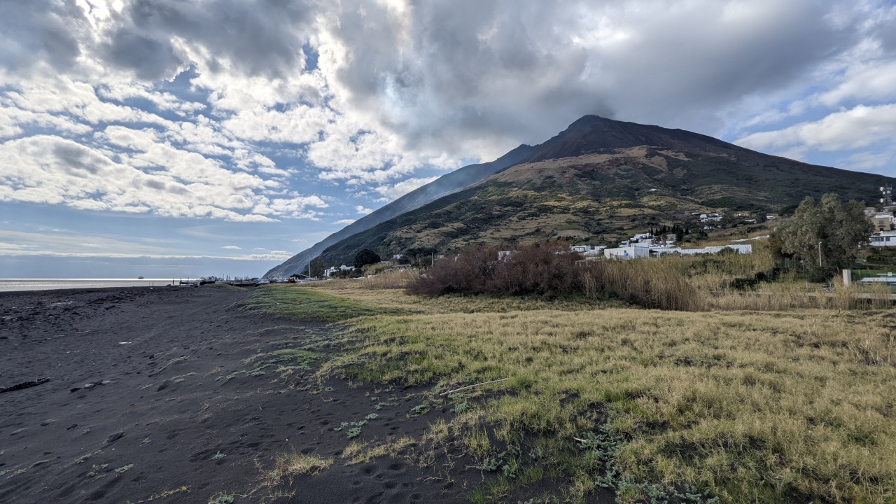 Die Insel Stromboli, im Hintergrund ist der Vulkan mit dem Dorf zu sehen. Im Vordergrund ist schwarzer Sand als Strand. 