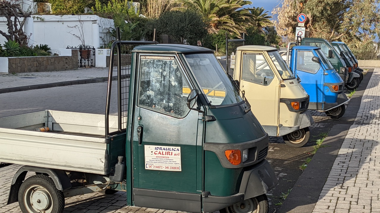 Mehrere dreirädrige Ape Motorroller mit Ladefläche parken am Strassenrand. 