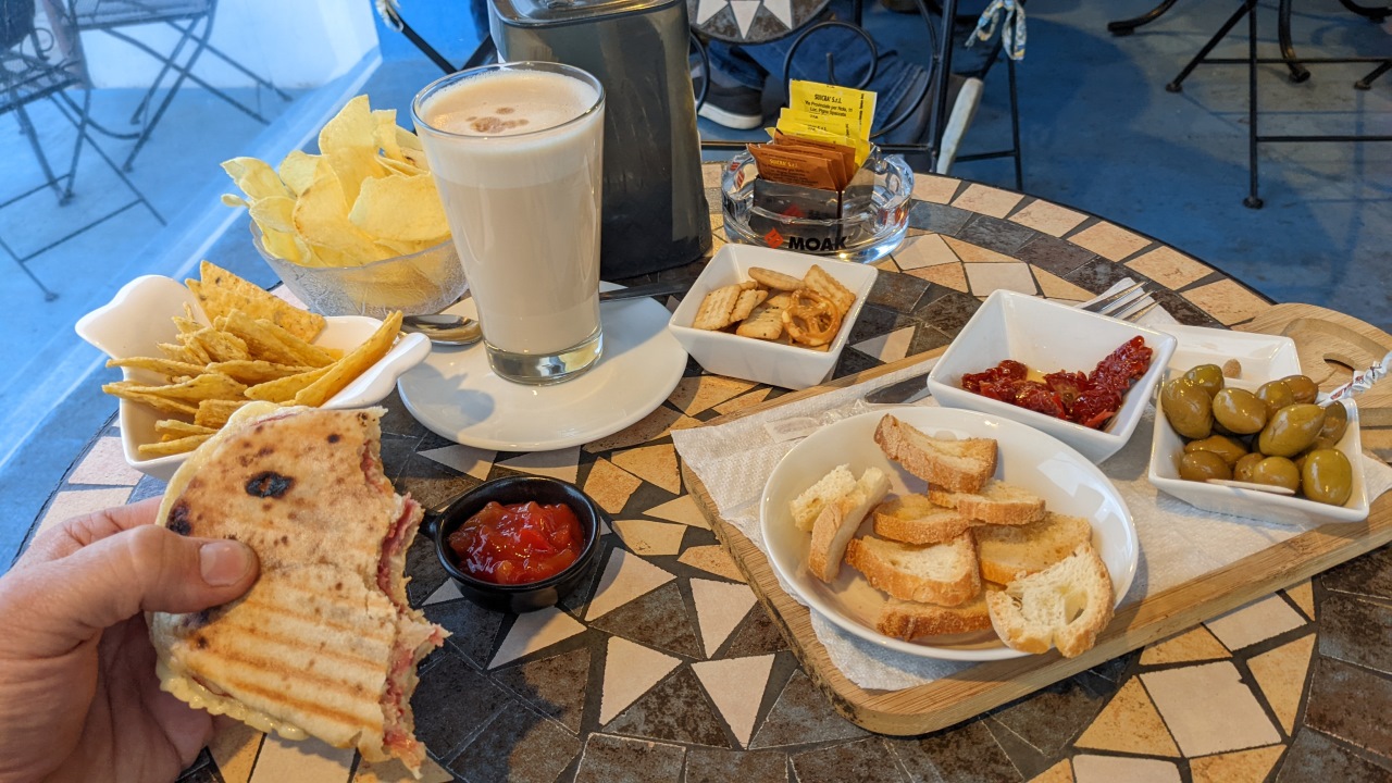 Eine Auswahl von Snacks im Blu Bar Cafe: Chips, Tacos, Oliven, gebackenes Brot, Caffe Latte, Sandwich, eingelegte Tomaten, rote Sauce