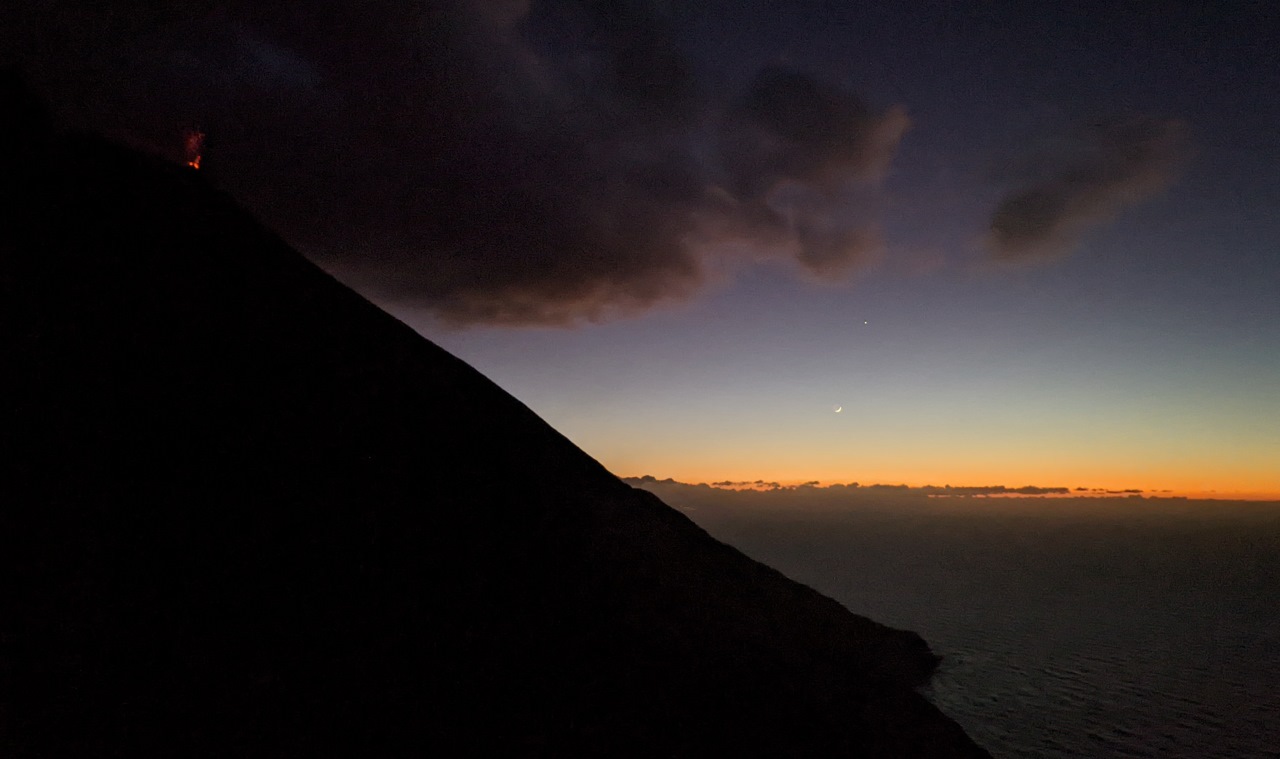 Der Vulkan in der Abenddämmerung  fotografiert. Der Berg ist komplett schwarz, oben rechts ist ein kleiner roter Feuerschein von einem Vulkanausbruch. Die Sonne ist unter gegangen, am dunkelblauen Himmel sind Wolken, die dünne Mondsichel und der hell leuchtende Jupiter zu sehen. 
