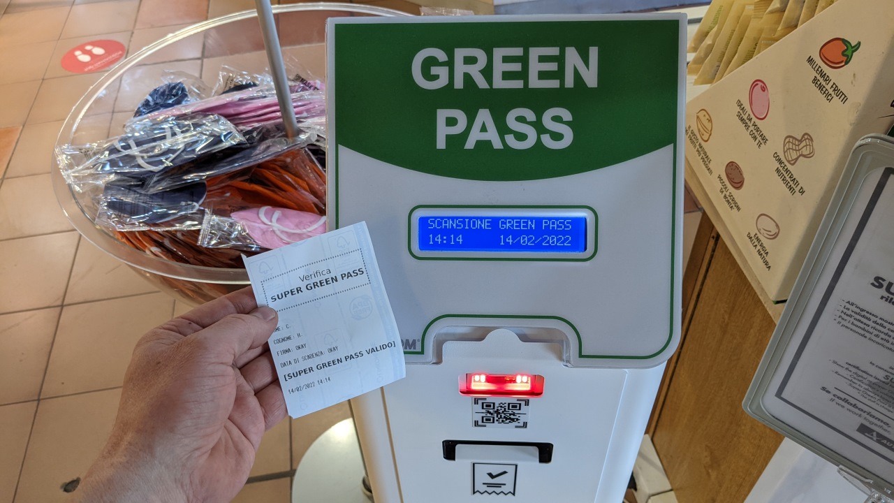 Ein italienischer Automat, der die Green Pass App zur Dokumentation der Corona Impfung auslesen kann und dann einen Papier Beleg druckt. Ich halte einen solchen Beleg ins Bild, vor dem im Hintergrund stehenden Automaten. 
