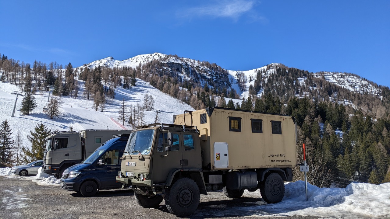 Der DAF T244 Truck parkt vor einem blauen MAN Camper Van und einem 16 Tonnen Expeditionsmobil. Im Hintergrund vom Parkplatz sind verschneite Berge und der blaue Himmel zu sehen. 