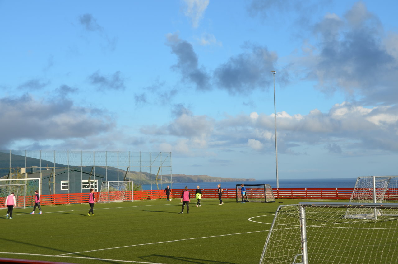 Ein Fussballplatz auf dem mehrere Frauen trainieren. Der grüne Fussballplatz ist höher gelegen, im Hintergrund ist unten das blaue Meer zu sehen. Der Himmel ist blau mit dunklen Wolkenfetzen. 