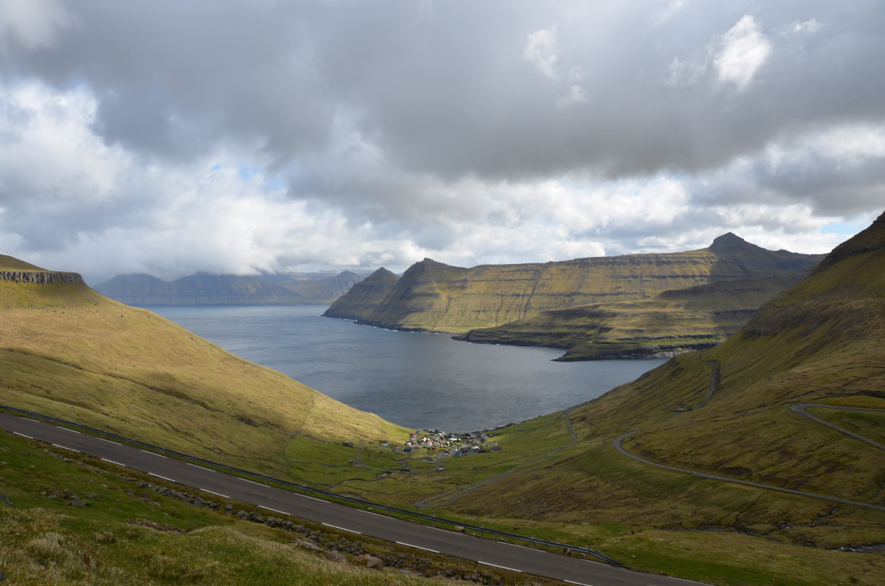 Sundini Fjord Färöer Inseln, die Sonne scheint schräg auf die Berge. Der Himmel ist wolkenbedeckt. Unten am Wasser liegt ein kleines Dorf. 