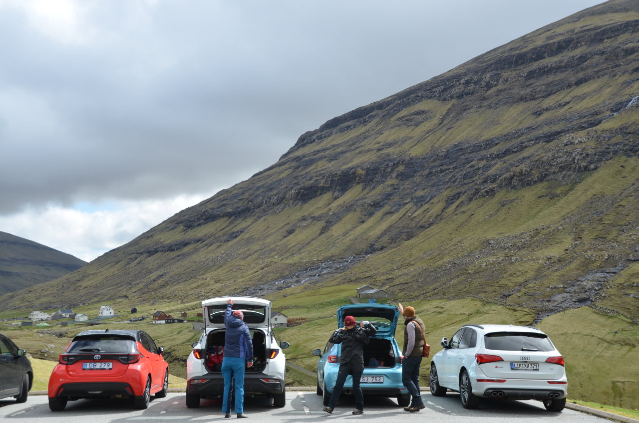 Vorne stehen vier Autos im Tag von Saksun, zwei davon haben die Heckklappe geöffnet. Drei Personen stehen bei den Fahrzeugen. Im Hintergrund ist ein grünes Bergmassiv. 