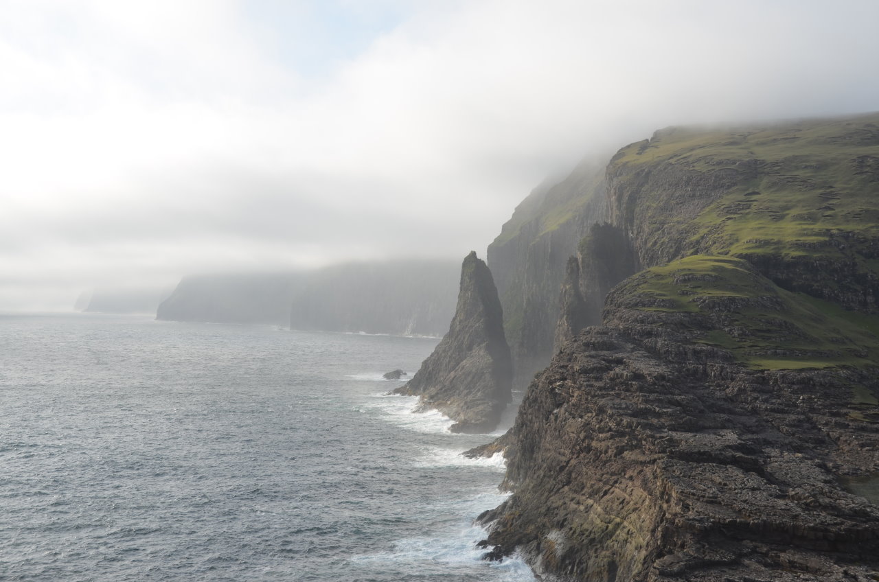 Blick auf die zerklüftete Küste beim Bosdalafossur. Links ist das blaue Meer mit etwas Nebel, rechts Gebirge mit grünen Gradflächen. 