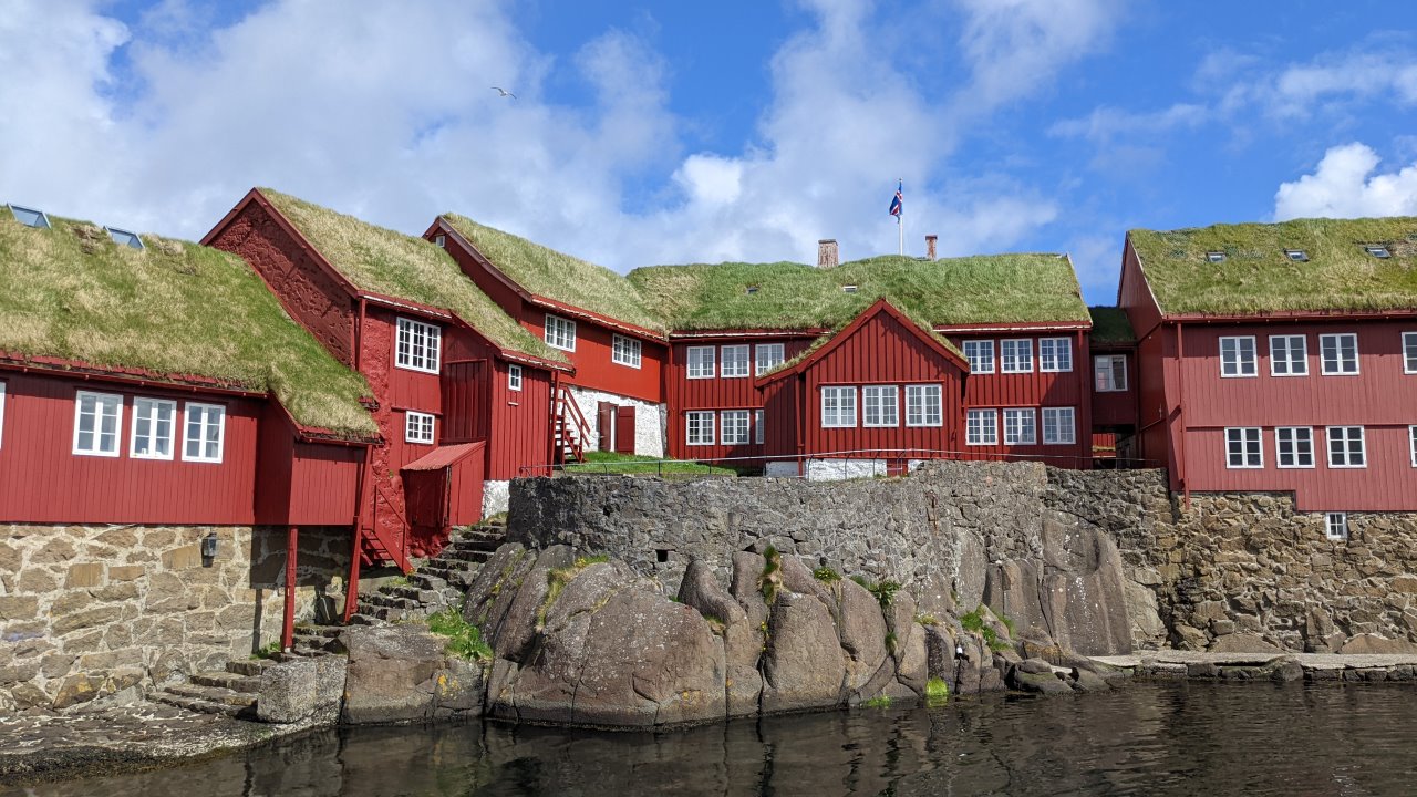 Eine Reihe rot angemalte Holzhäuser mit grünen Grasdächern und weissen Fenstern stehen auf auf Felsen. Im Vordergrund ist etwas dunkles Wasser zu sehen. 