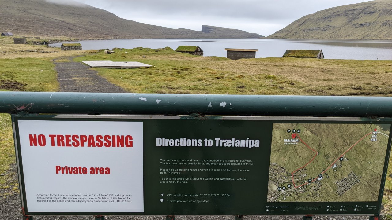 Der Vordergrund zeigt an einer Absperrung ein Hinweisschild, welches Touristen auf Englisch den Weg zu einer Bezahlstation weist. Die Wörter no trespassing und private area sind rot hervor gehoben. Rechts auf dem Schild ist eine Luftaufnahme abgebildet, wo der Weg zum offiziellen Eingang mit der  Bezahlstation rot eingezeichnet ist. Obwohl das Schild sehr viel erklärenden Text und eine Drohung enthält fehlt die wichtige Information  wie viel bezahlt werden muss. Im Hintergrund ist der See Leitisvatn zu sehen. Am Ufer stehen drei bis vier alte Fischerhütten mit Grasdach.  