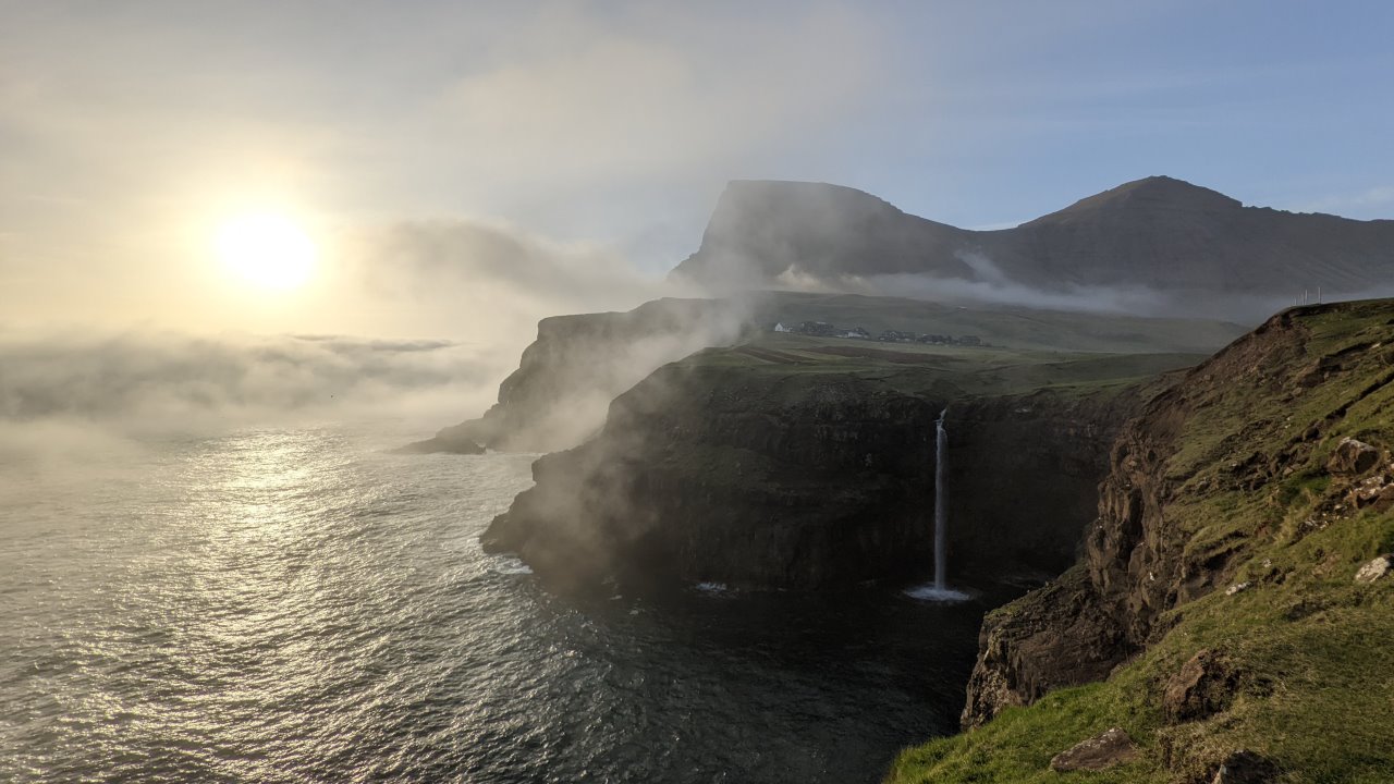Sonnenuntergang bei Gasadalur. Der  Wasserfall ist rechts im Bild und fällt tief hinab ins Meer. Die Sonne scheint durch Wolkenfetzen hindurch, fotografiert auf den Färöer Inseln