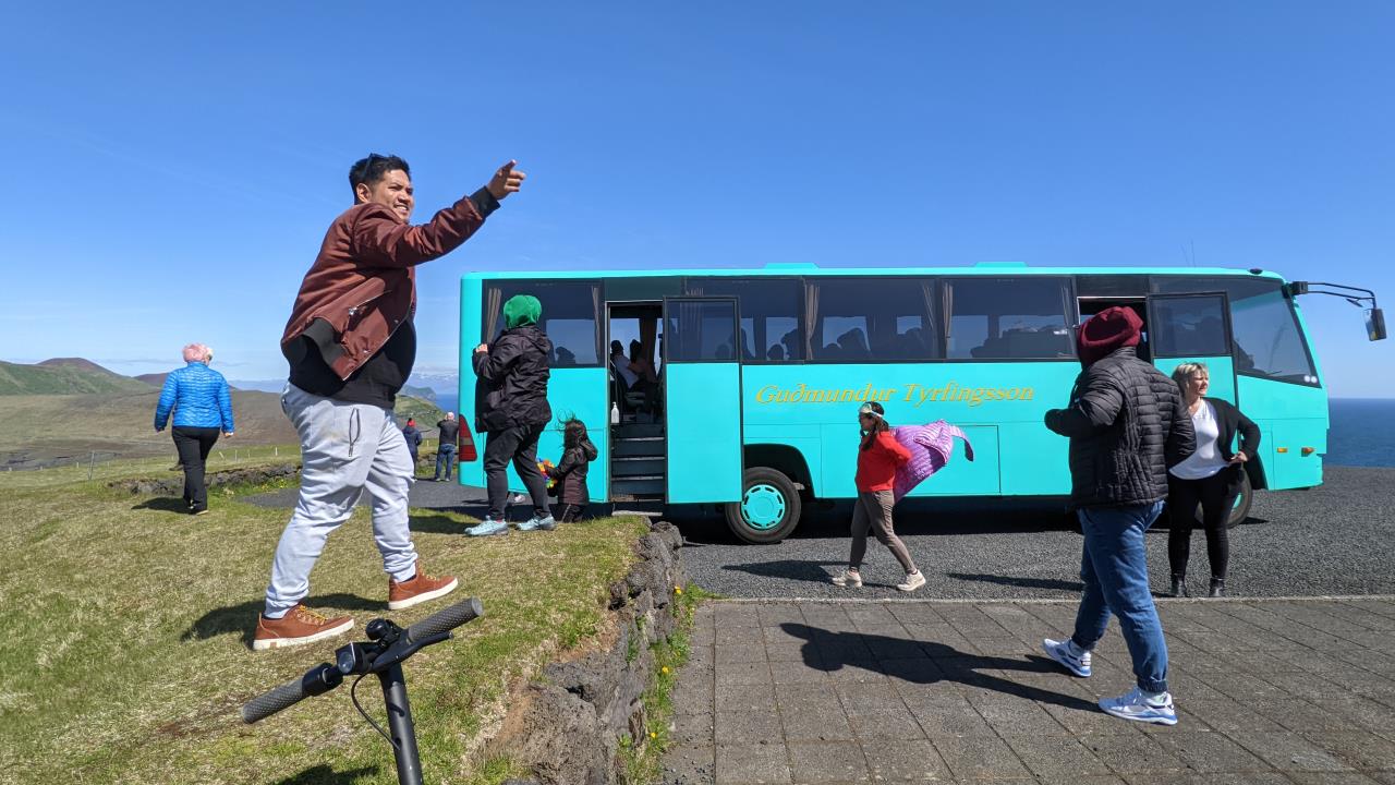 Türkiser Reisebus, aus dem neun Touristen ausgestiegen sind, Diese laufen bei starkem Wind um den Bus herum. Ein Tourist asiatischer Abstammung zeigt begeistert auf etwas. 