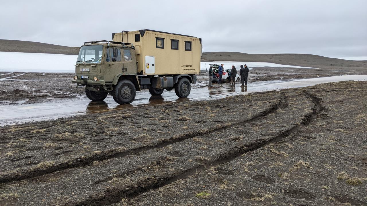 Ein DAF T244 Gelände LKW steht sicher auf einer Piste in Island. Im Vordergrund sind Fahrzeugspuren im Matsch zu sehen. Hinten rechts hat sich ein weisses Fahrzeug im Matsch festgefahren, um das fünf Personen herumstehen. 