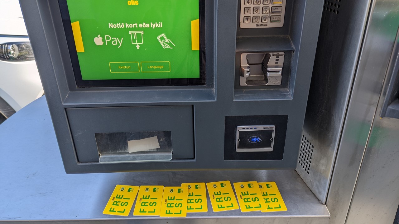 Eine Olis Tanksäule mit grünem Bildschirm, Kartenleser und Zahlen Tastatur. Unter dem Ausgabefach für die Papier Quittung liegen 7 benutzte, gelbe Olis 5000 ISK Guthabenkarten aus Plastik.  