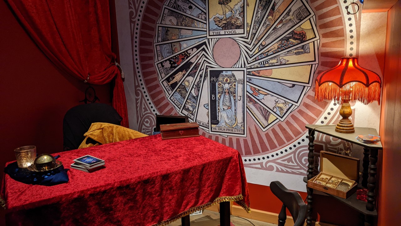 Tisch einer Wahrsagerin mit roter Tischdecke aus Samt. Ein Bild an der Wand zeigt Tarot Karten 