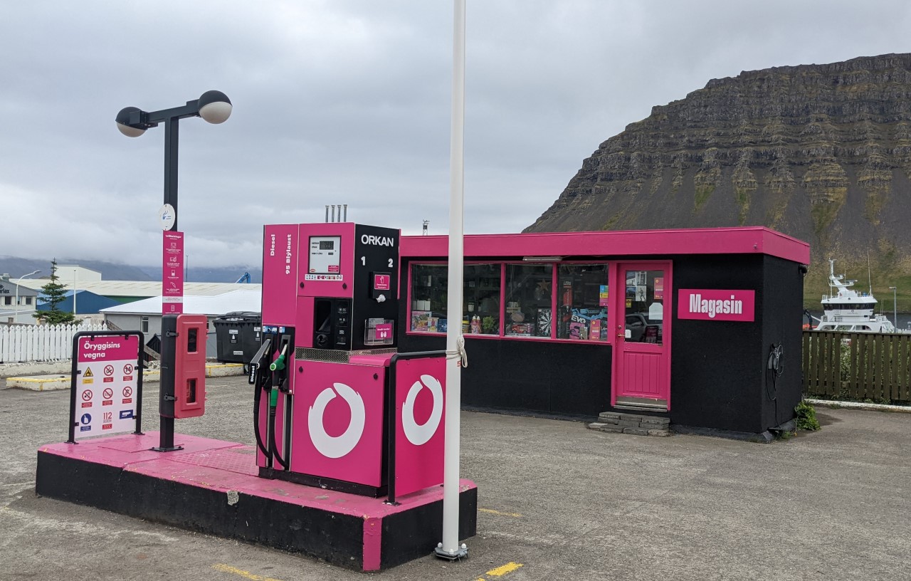 Eine Tankstelle in Island von Orkan mit pinken Tanksäulen und Shop. Der Shop hat ein pinkes Dach und ein Schild mit Magasin. Im Hintergrund sind Berge. 