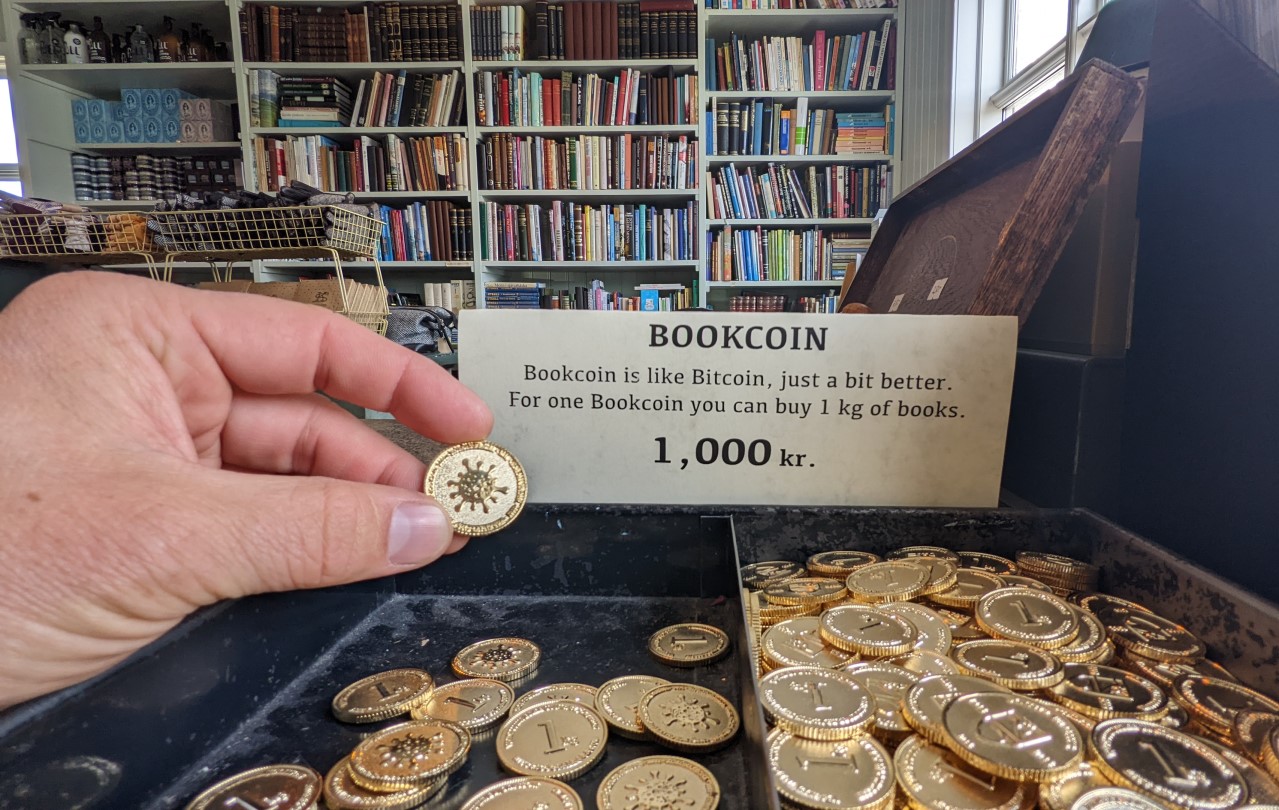 Eine Hand hält in einem Buchladen eine goldenen Münze mit dem Corona logo hoch. Die Münze wird auf einem Schild als Bookcoin bezeichnet. 