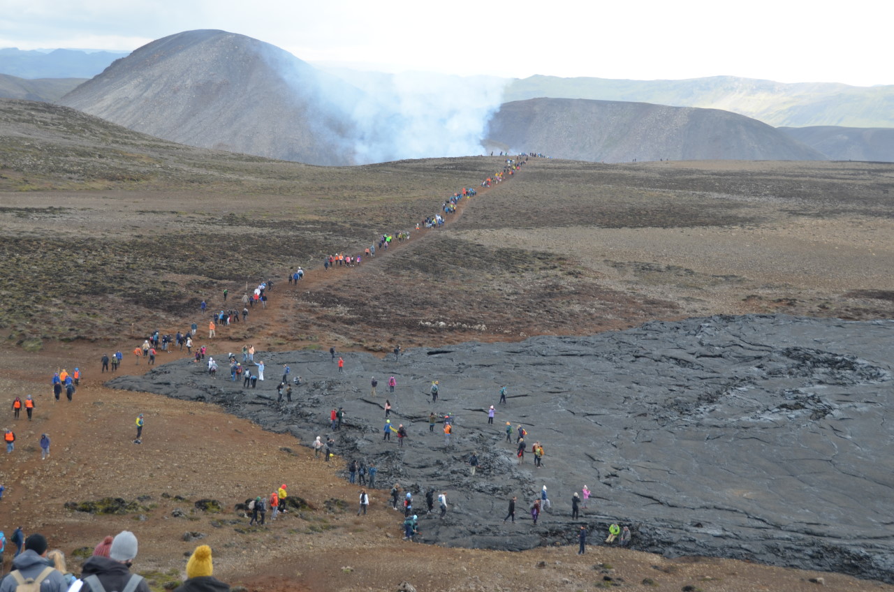 Menschen gehen auf einem Wanderweg zum Vulkan Vulkan Fagradalsfjall. In Vordergrund ist ein erkaltetes, schwarzes Lavafeld zu sehen, auf dem einige Menschen spazieren gehen. 