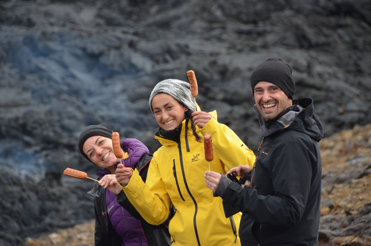 Eine Frau mit lila Weste und eine Frau mit gelber Regenjacke und ein Mann mit schwarzer Jacke haben Würstchen am Spiess auf der Lava gegrillt. Die drei Menschen halten die Würstchen fröhlich lachend hoch