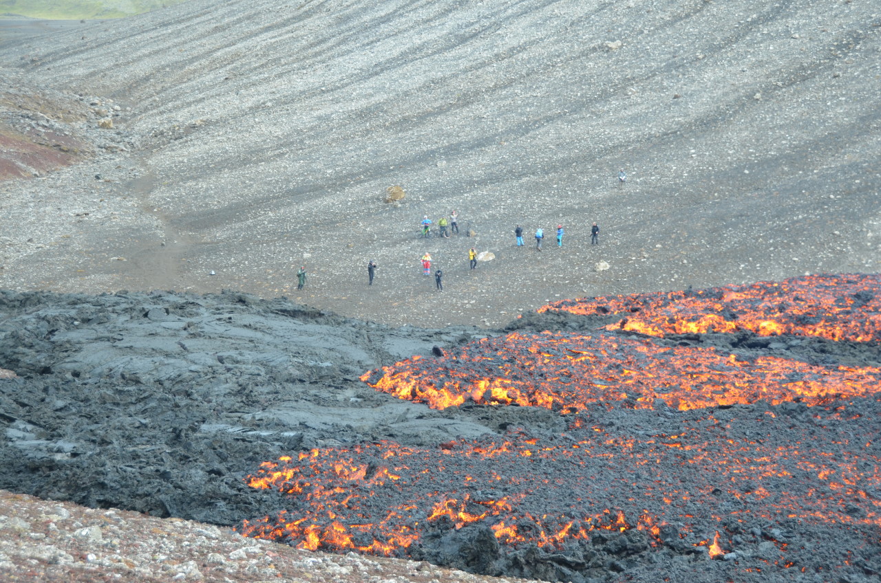 Menschen betrachten im Hintergrund eine rot glühende Lava Masse vom Meradalir Vulkan, die sich langsam voran bewegt. 