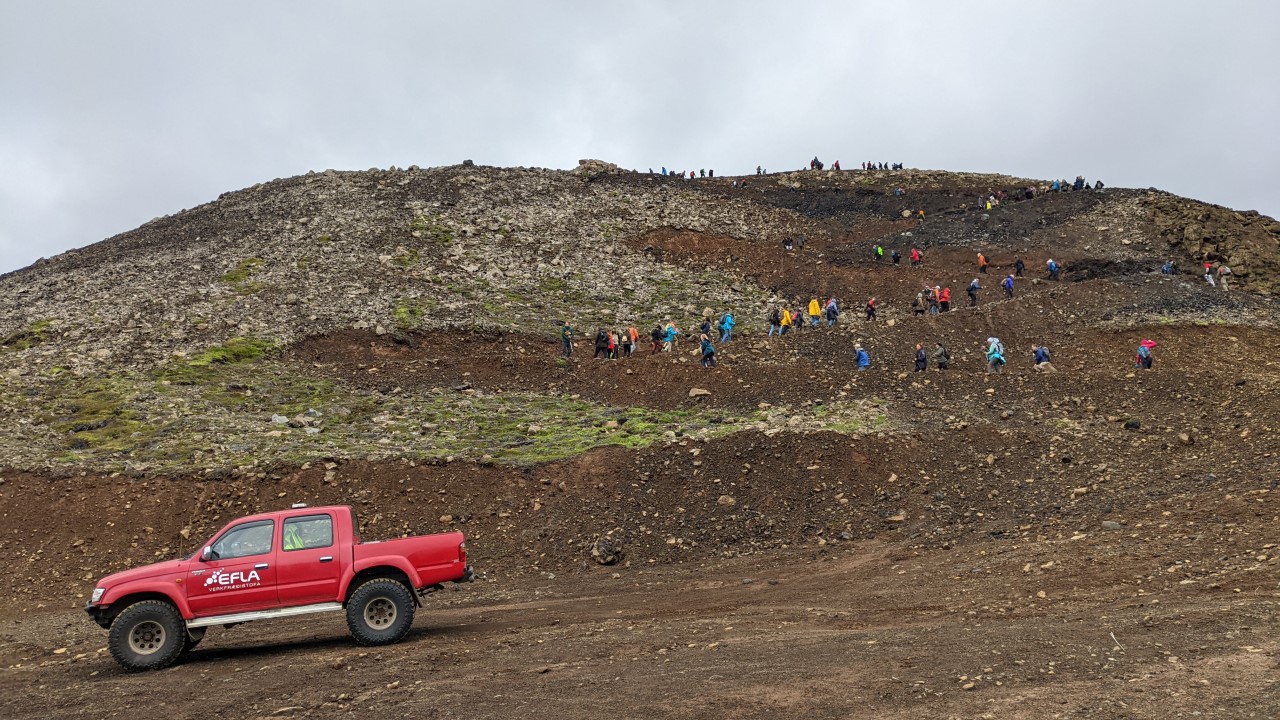 Eine grosse Gruppe menschen wandern im zick zack einen Berg hoch. Unten rechts steht ein roter Geländewagen. 