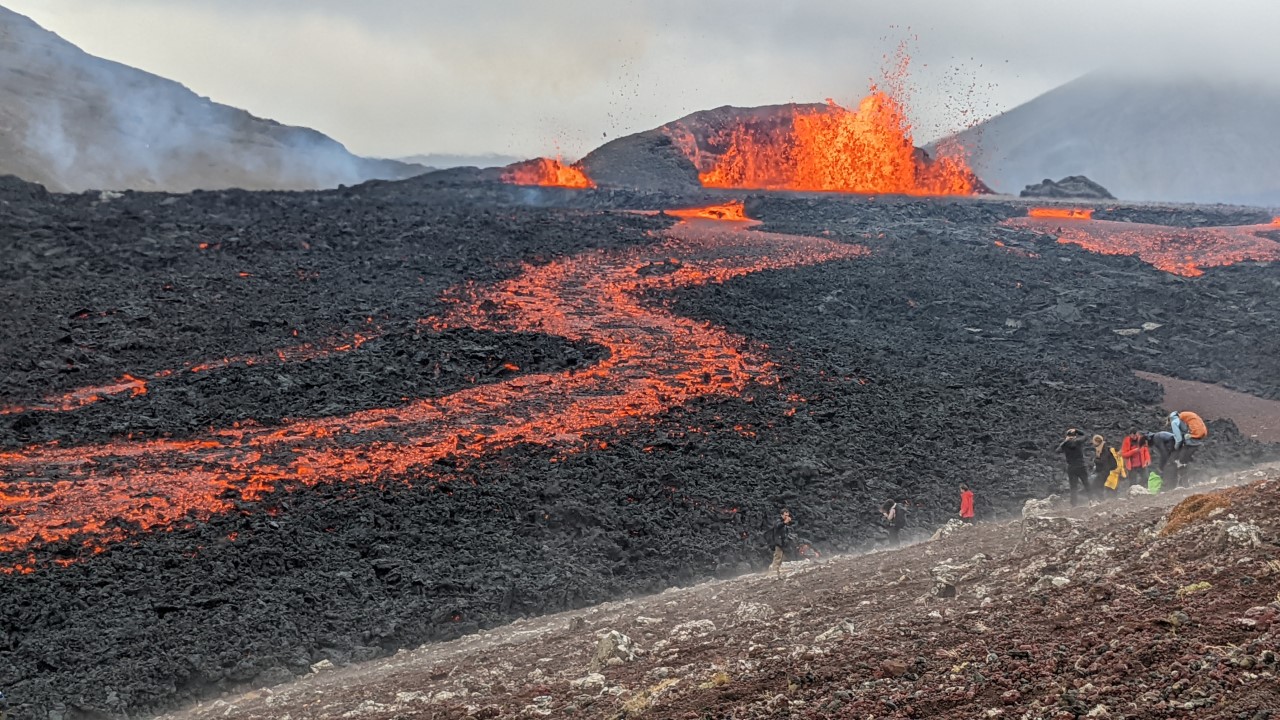 Vulkanausbruch vom Meradalir Vulkan Fagradalsfjall, der Lava spuckt die in ein Tal fliesst. Acht Menschen stehen rechts an einem Hang und beobachten die Eruption. 