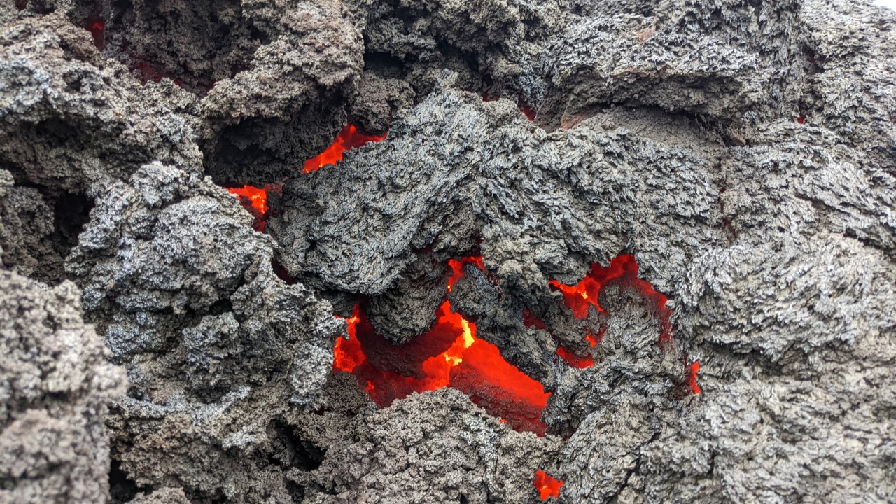 Nahaufnahme von grauer Lava, in einem Spalt glüht rot heisse Lava. 