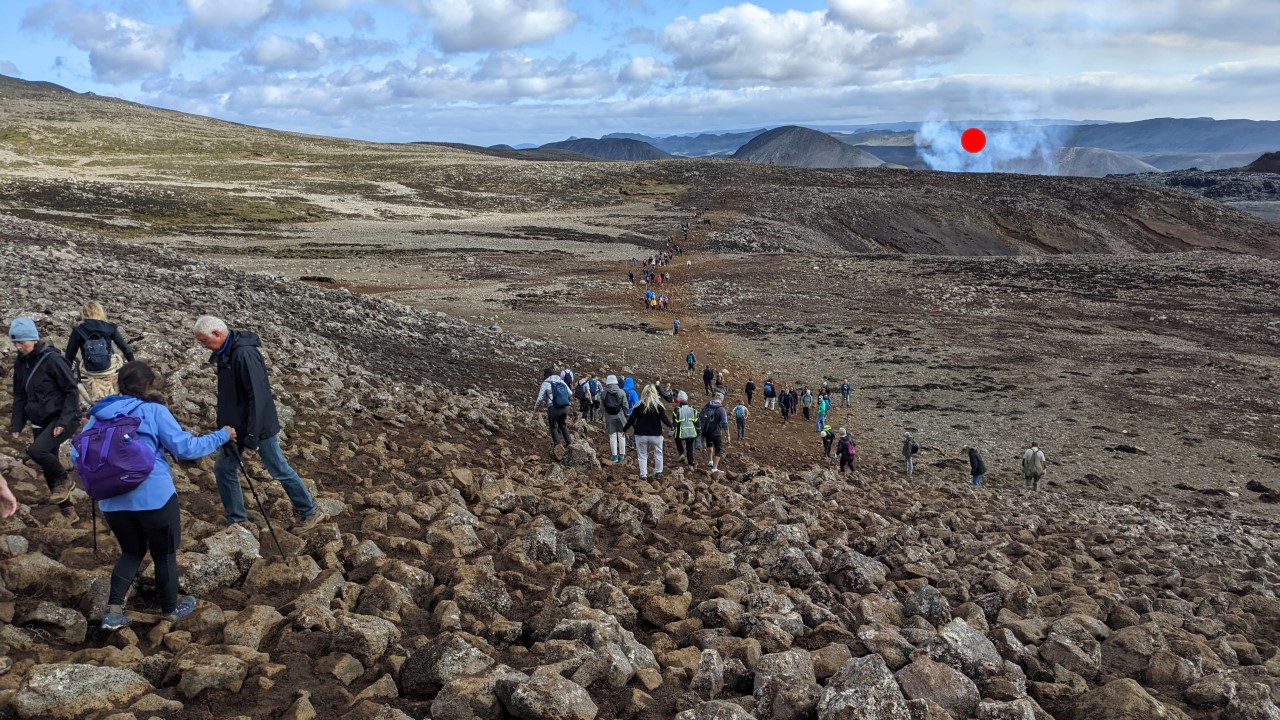 Wanderer sind auf sehr steinigem Untergrund auf dem Wanderweg  zum Vulkanausbruch Fagradalsfjall vom August 2022 unterwegs. Im Hintergrund ist die Rauchwolke vom Vulkan mit einem roten Punkt als Wegweiser markiert. 