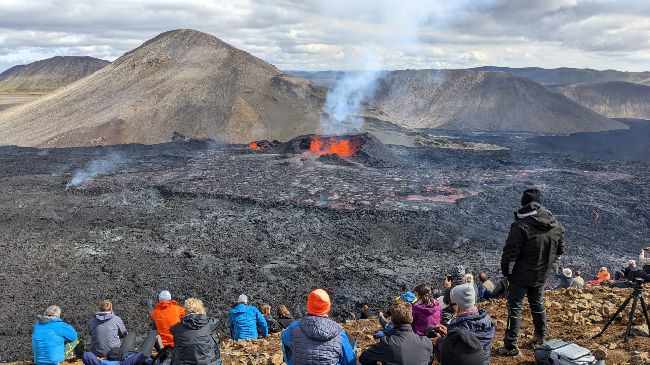 Zuschauer betrachten im August 2022 von einem Berg aus den Vulkanausbruch Fagradalsfjall. Der Vulkan spuckt glühende Lava, das Tal ist mit schwarzer erkalteter Lava bedeckt.   