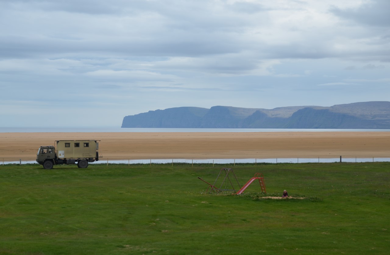 Ein LKW parkt auf einer grünen Wiese mit einem Kinder Klettergerüst und Rutsche. Im Hintergrund ist ein Sandstrand und Berge. 