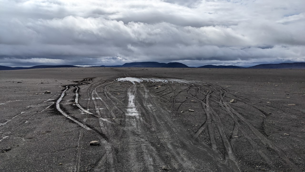 Island Hochland Panorama mit grauem Boden und grauen Wolken. In der Mitte vom Bild ist eine Piste zu erkennen, die durch eine kleine Stelle mit Wasser führt. Sehr viele Auto Spuren führen durch den Matsch links und rechts daran vorbei. 