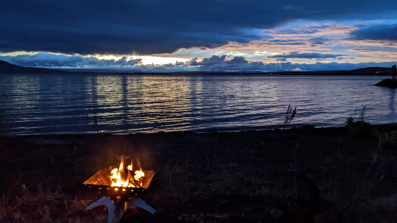 Lagerfeuer in einer Feuerschale am dunklen Strand am Myvatn See Island. Wolken und ein Rest Tageslicht spiegeln sich im See. 