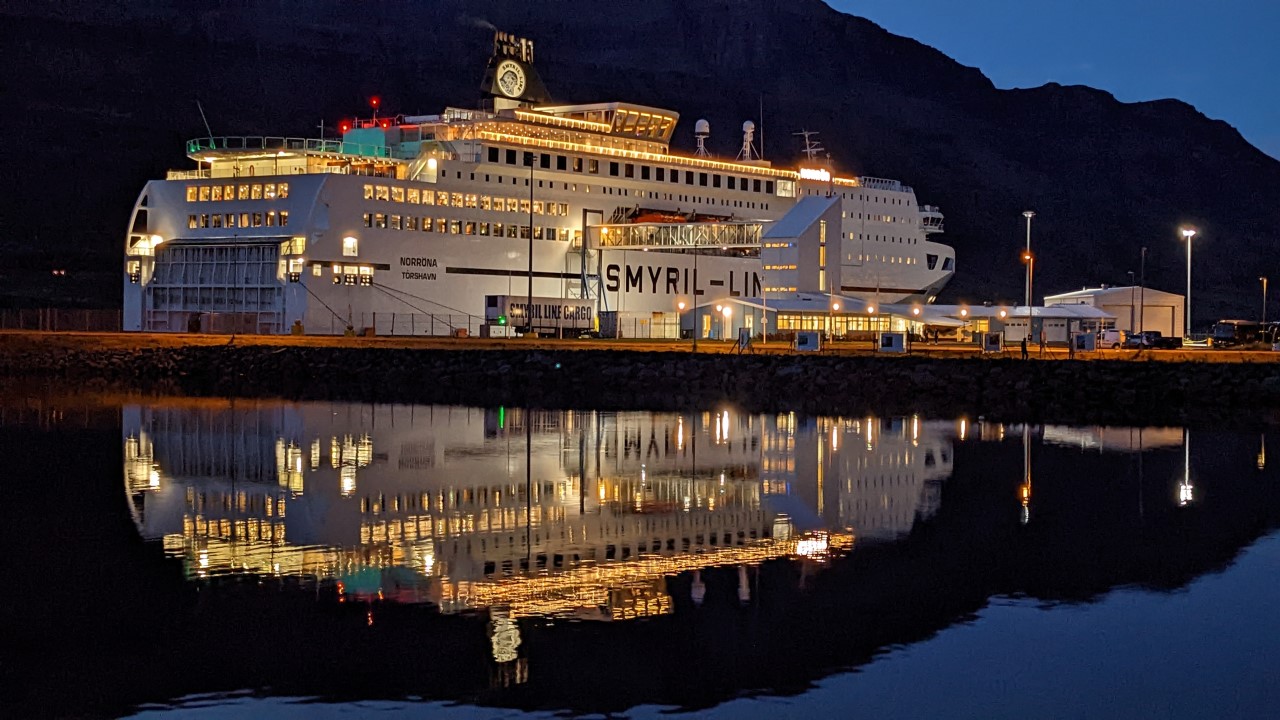 Nachtaufnahme der Norröna Fähre im Hafen von Seydisfjördur. Die hell beleuchtete Fähre spiegelt sich im Hafenbecken. 