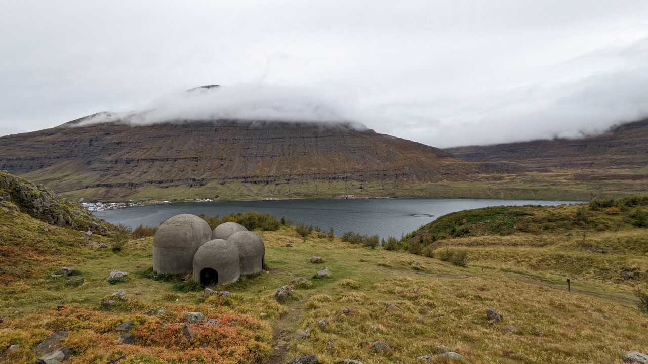 Vier runde Betonhäuschen stehen am Rand von Seydisfjördur Fjord. Die Berge im Hintergrund sind Wolkenverhangen. 