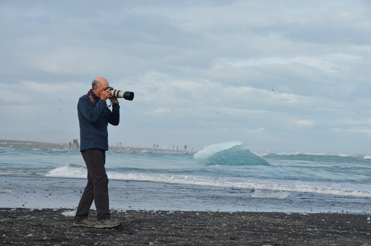 Ein Mann läuft mit einer Kamera mit ausgefahrenem Zoom Objektiv vor dem Gesicht am Diamond Beach  Jökulsarlon Island entlang. 