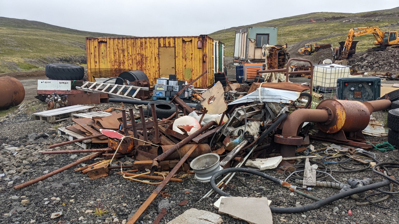 Schrottplatz in Island mit rostigen Metallteilen, Blei Batterien, Fässern und aufgegebenen Baumaschinen und einem gelben Container. 