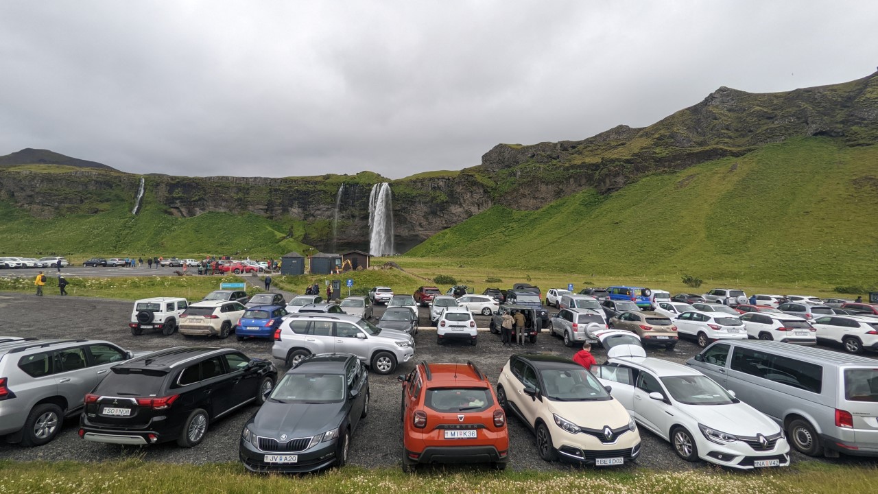 Parkplatz mit sehr vielen Autos vor einem Wasserfall in Island