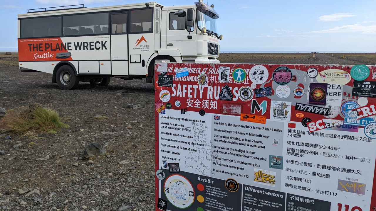 Ein Touristenbus, der mit The Plane Wreck Shuttle beschriftet ist parkt hinter einen Übersichtsschild. Auf dem hinteren, linken Reifen hat der Bus zu wenig Luft. Das Schild mit Hinweisen in Isländisch, Englisch und Chinesisch ist mit zahlreichen bunten Aufklebern beklebt.  