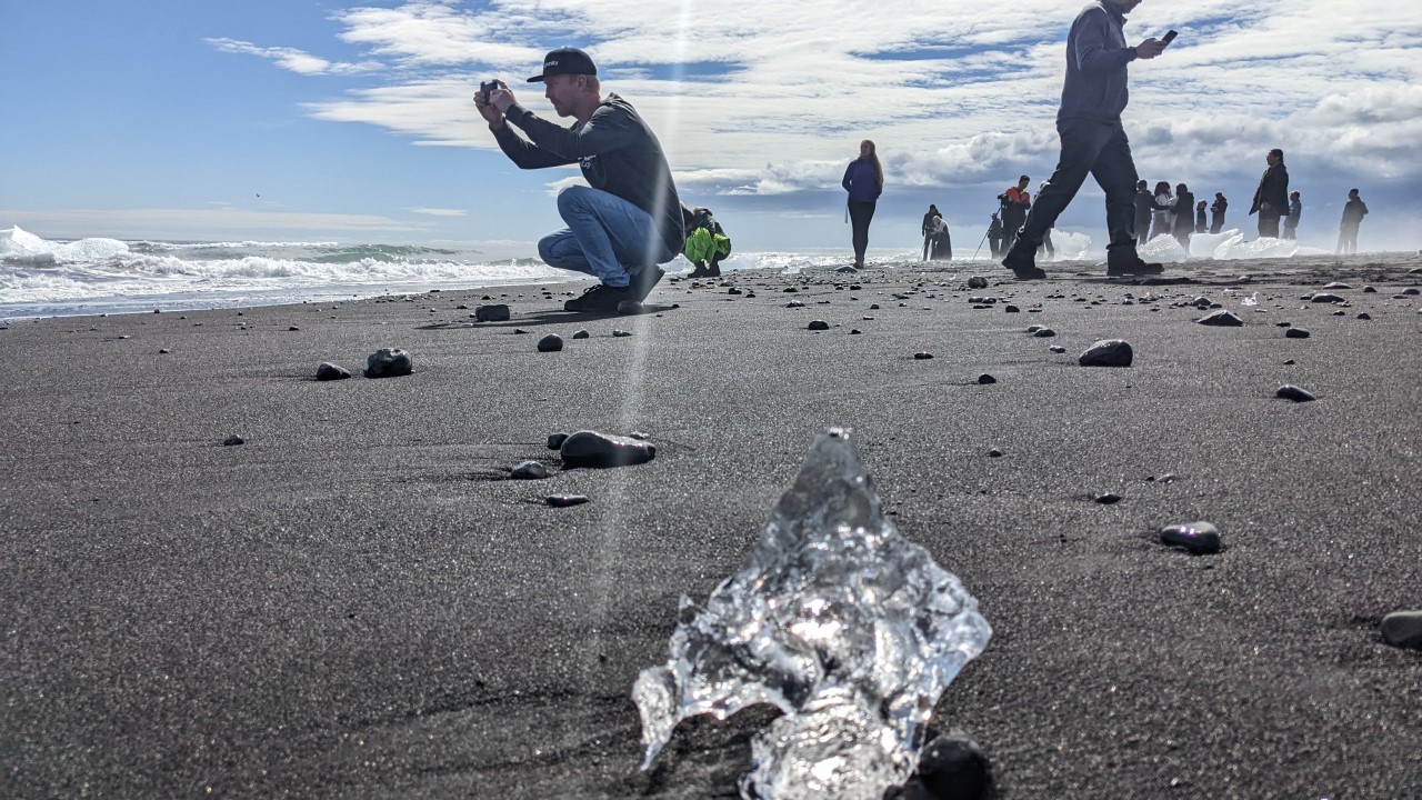 Diamond Beach Strand am Jökulsarlon in Island mit fotografierenden Touristen und angeschwemmten Eisbrocken. Im Hintergrund ist das Meer und blauer, wolkiger Himmel. 