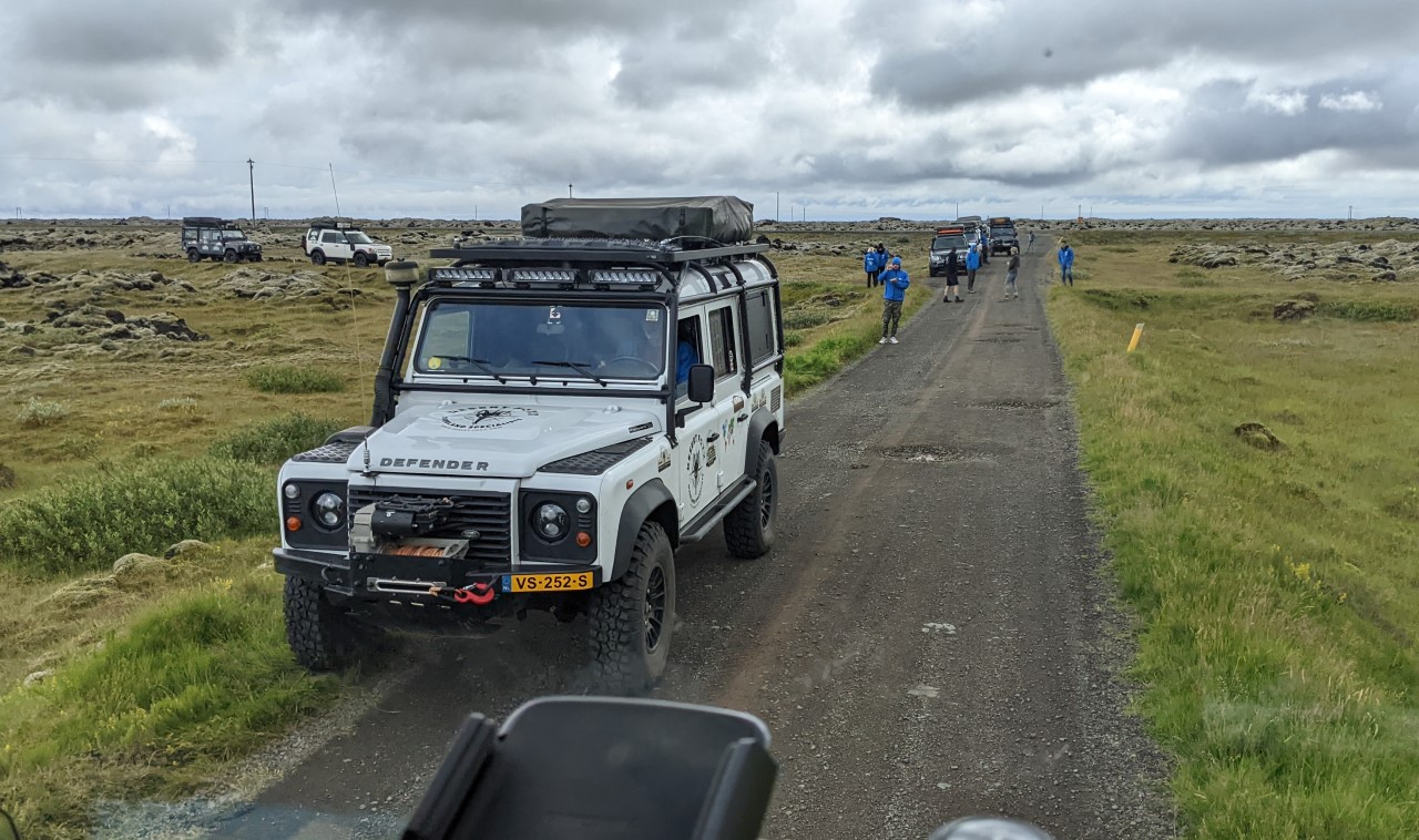 Sieben Landrover Geländewagen auf einer grauen Piste, um die Fahrzeuge laufen Personen herum. 