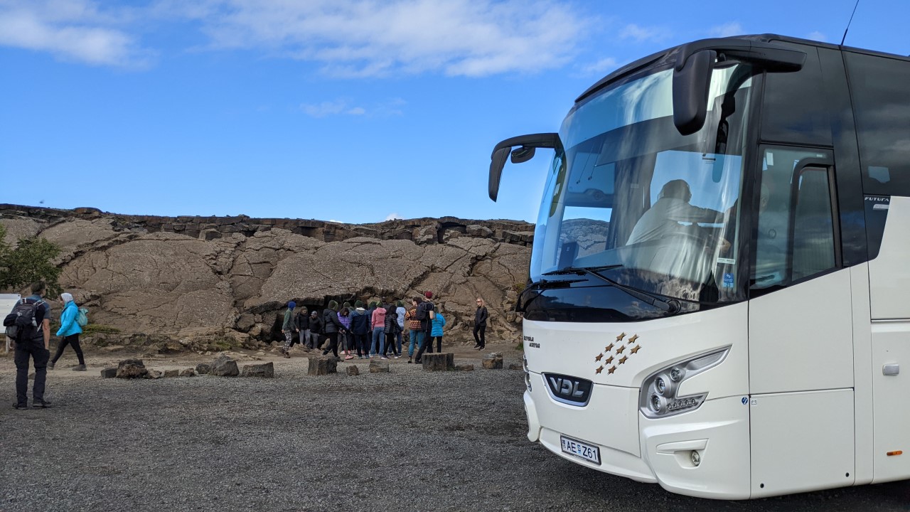 Eine Reisegruppe steht vor der Grjotagja Höhle in Island Schlange, um diese zu besuchen. Rechts parkt ein weisser Reisebus von VDL. 