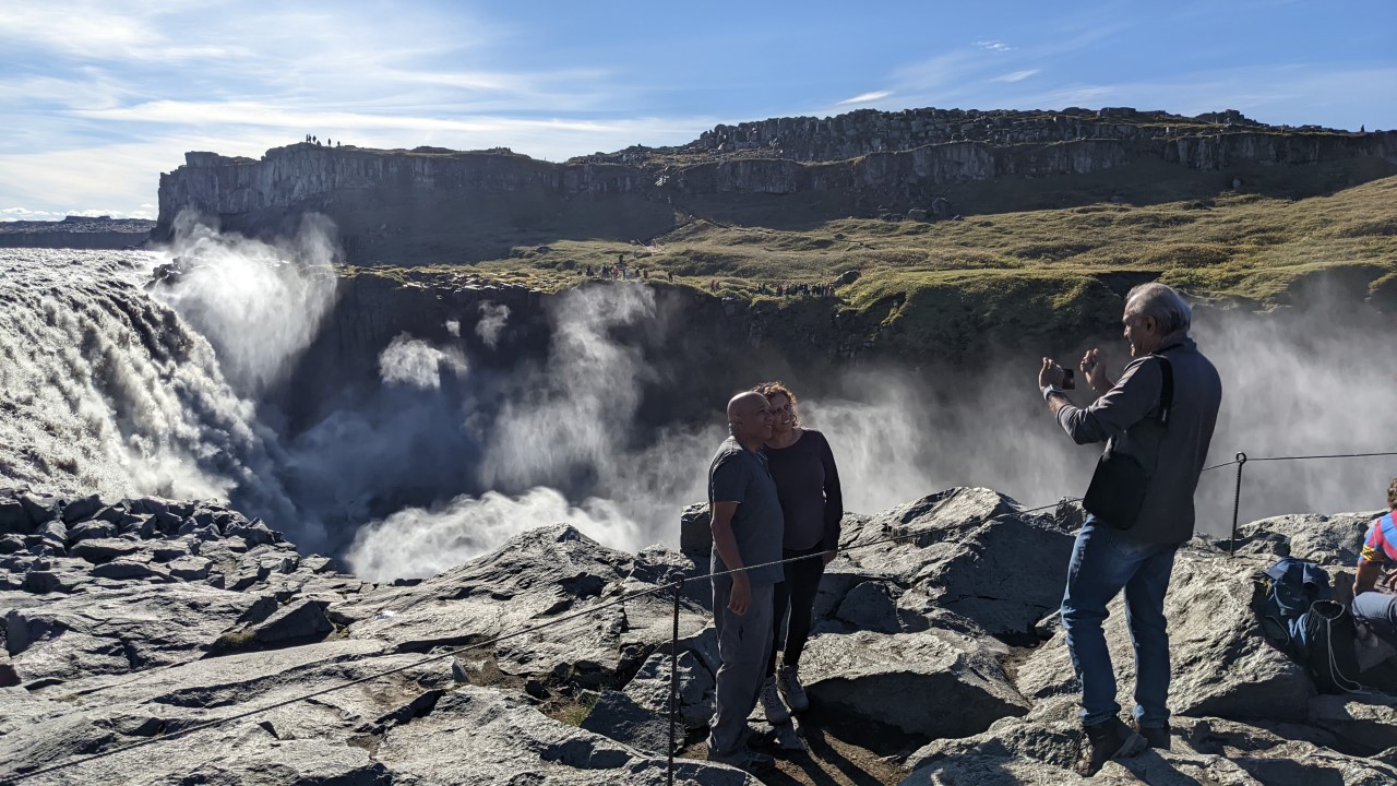 Ein Pärchen steht hinter einer Absperrung aus einem Seil und wird von einer dritten Person fotografiert. Im Hintergrund ist der Dettifoss Wasserfall in Island. 